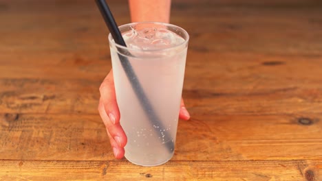 Primer-Plano-De-Un-Refrescante-Vaso-De-Agua-Mineral-Con-Hielo,-Una-Pajita-Y-Un-Toque-De-Limón-Que-Se-Quita-De-Una-Mesa-De-Madera-En-Un-Bar