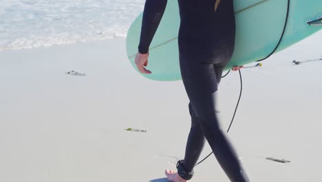 video von einem kaukasischen mann in wetsuit mit einem surfbrett, der auf einem sonnigen strand ins meer geht