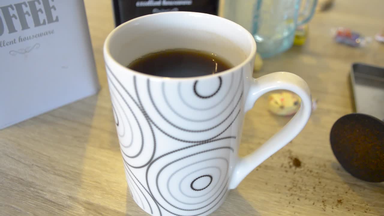 белая кружка, полная горячего черного кофе, сидящая на столе с контейнерами для кофе, сладостями и винтажным стеклом, куколка в