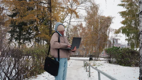 light skin lady standing outdoors on snowy path wearing warm sweater jacket, holding laptop and talking on phone while surrounded by autumn trees and residential buildings in soft natural light