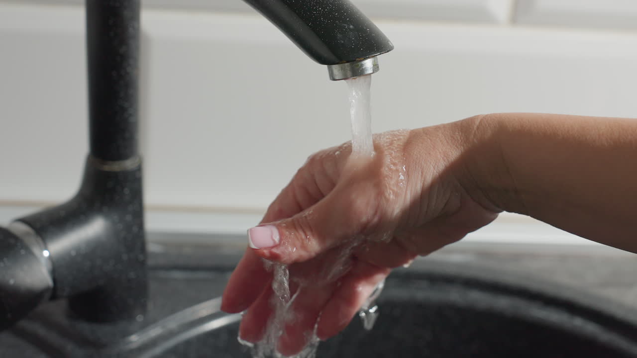 Close up of female hand with light pink nails rinsing under flowing kitchen tap water, visible soap bubbles forming on skin, emphasizing hand hygiene and personal care