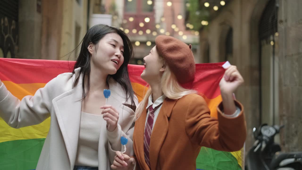 Two women celebrate LGBTQ pride with a rainbow flag and lollipops on a city street