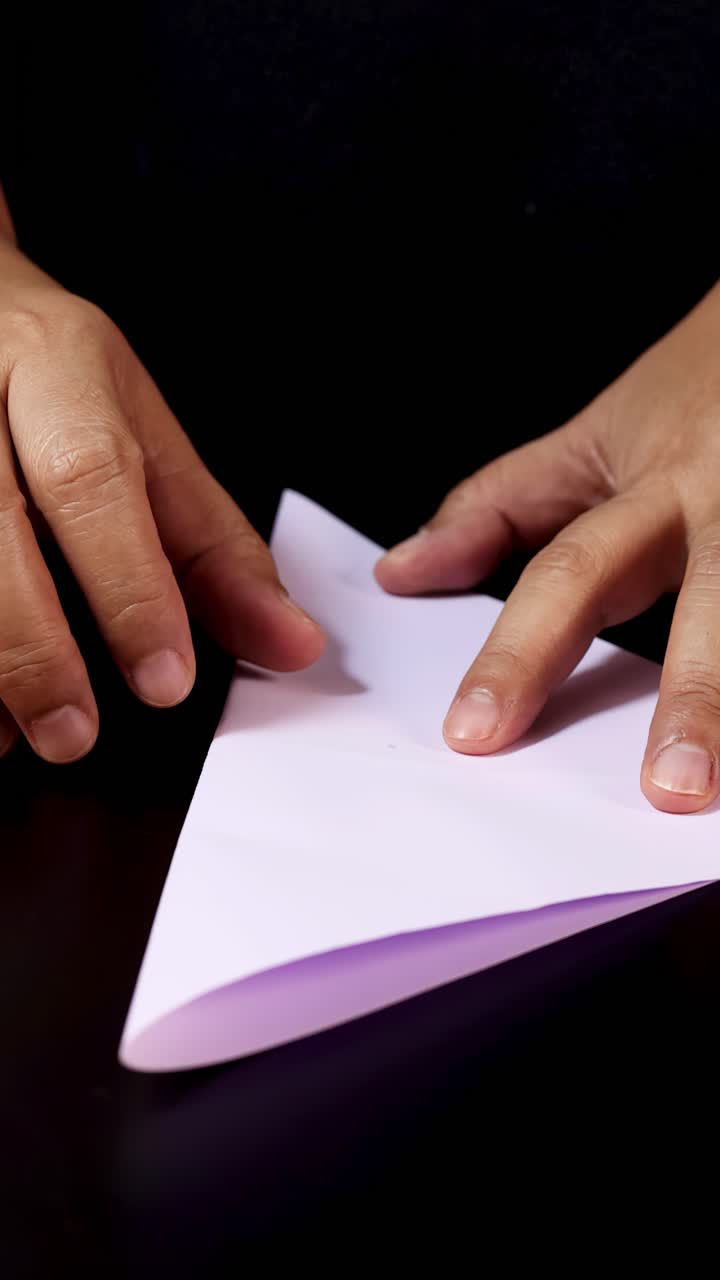 Mani adulte piegano con cura carta bianca su una superficie nera sotto un'illuminazione morbida e uniforme, vista ravvicinata