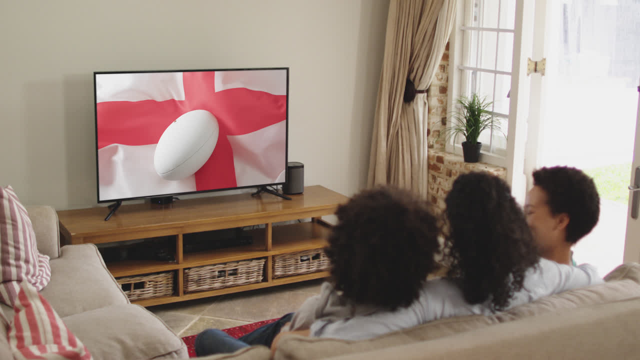Famiglia birazziale che guarda la TV con un pallone da rugby sulla bandiera dell'Inghilterra sullo schermo.