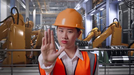 close up of asian female engineer with safety helmet disapproving with no hand sign while standing with industrial robotic arms working