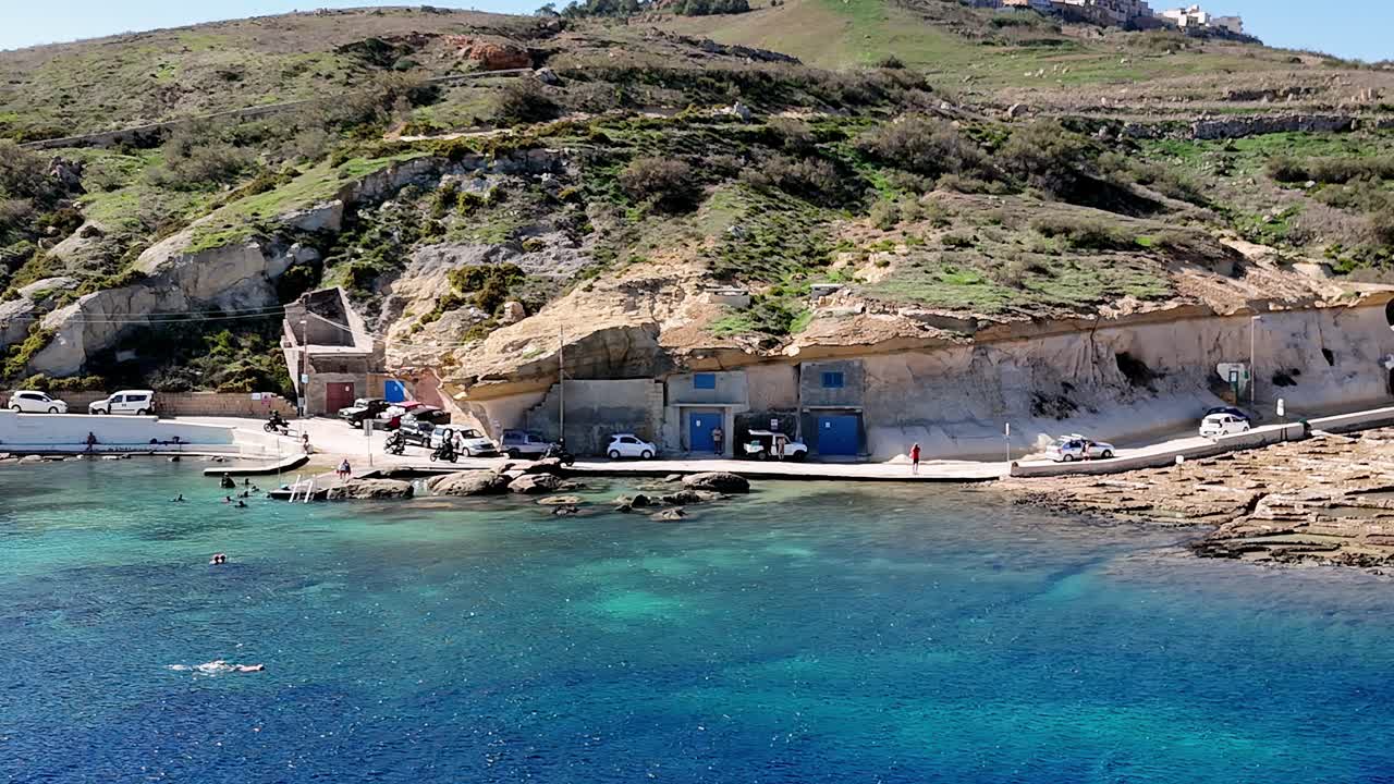 Eftermiddag ved Daħlet Qorrot Bay Gozo, Malta, med rustikke kalkstensbådhuse med livlige blå døre, der er hugget ind i de robuste klipper med udsigt over et krystalklart, glitrende turkisblåt Middelhav.