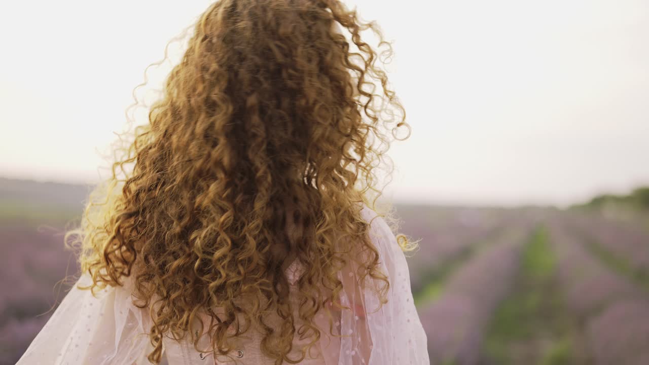 vista trasera de una mujer en vestido sosteniendo una cesta con algunos alimentos y flores de lavanda y caminando a través de un campo de lavanda
