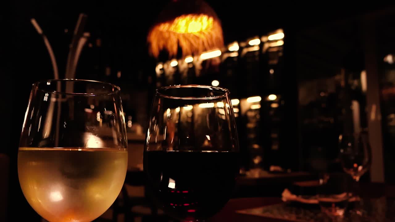 Órbita lenta ao redor de taças de vinho branco e tinto brilhantes com luzes bokeh quentes e plano de fundo de restaurante borrado, criando uma atmosfera aconchegante de jantar noturno em Lisboa