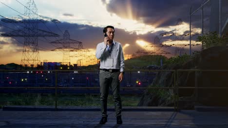 full body of an asian male professional worker standing near high voltage tower, industrial facility, he is speaking with someone in the phone
