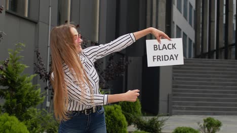 Happy-girl-holding-Black-Friday-inscription-text-and-pointing-on-it.-Shopping-mall-background