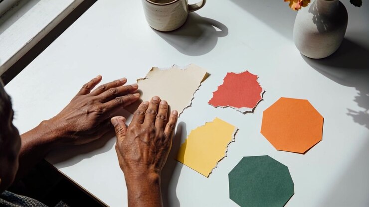 Entering left, hands in dark knit sleeve smoothing torn papers by window, making craft with shapes
