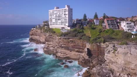 Disparo-Inverso-De-Drones-De-Olas-Oceánicas-Chocando-Contra-Rocas-Con-Agua-Turquesa-Y-Casas-De-Apartamentos-En-Un-Acantilado-En-El-Suburbio-De-Sydney,-Diamond-Bay-Australia