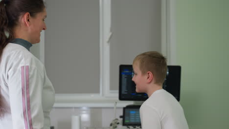 female pediatrician warmly welcomes child with smiles inside clinic, encouraging him to sit and lie down on examination bed while preparing for routine checkup in friendly, caring environment