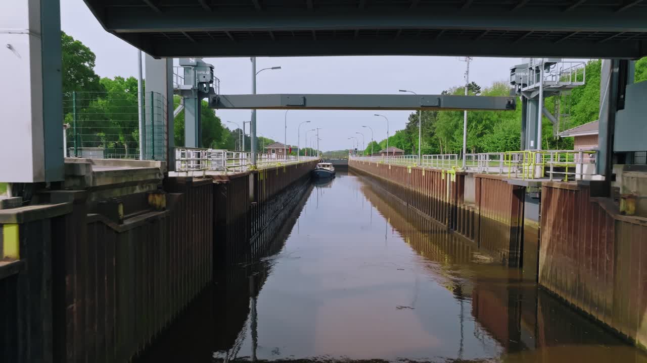 Zoom in to a white boat as it progresses through a canal lock with metal walls and bridges under soft morning sun and blue sky.