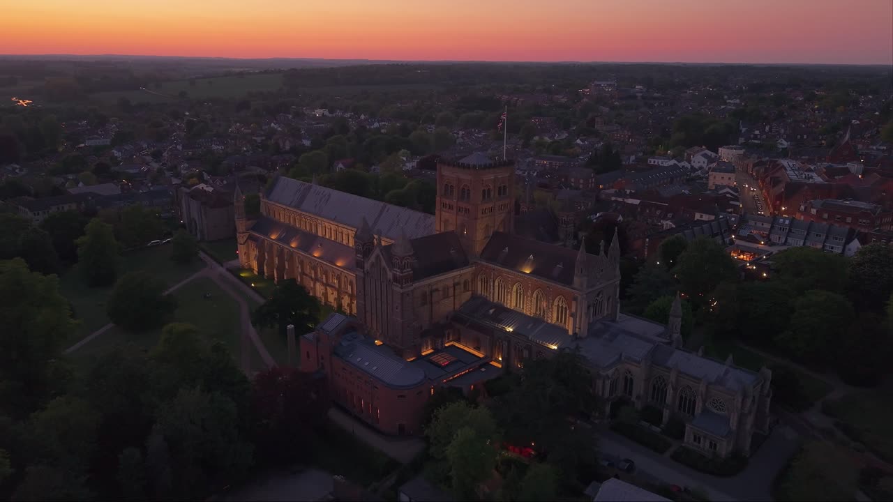 Aerial drone pullback captures St Albans Cathedral as the camera tilts up, revealing a stunning sunset over the city and casting warm light on the historic architecture