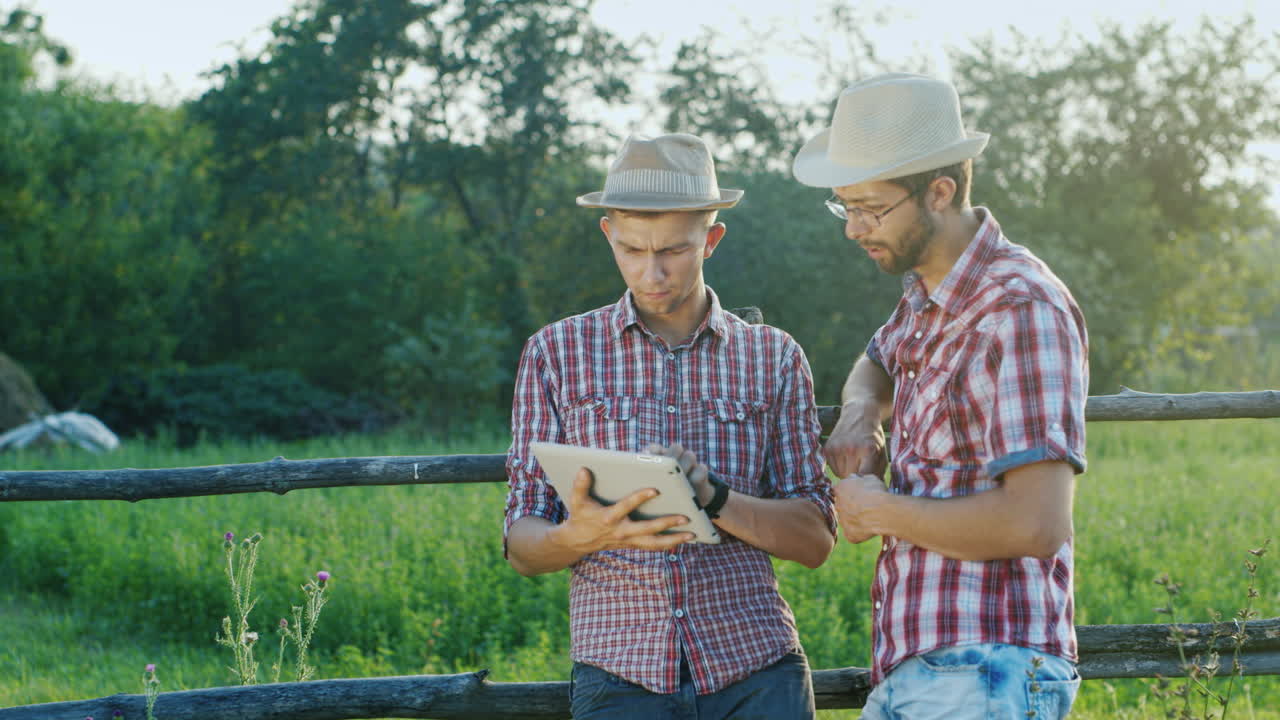 dos granjeros conversan en la cerca de un rancho y usan una tableta
