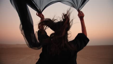 tracking-shot-of-an-Arabic-girl-holding-a-flying-black-scarf-while-running-in-the-desert-barefoot-at-the-fossil-dunes