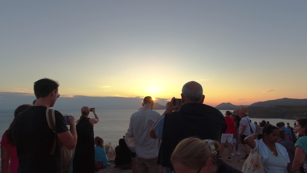 People standing on top of cliff overlooking the ocean taking pictures at sunset