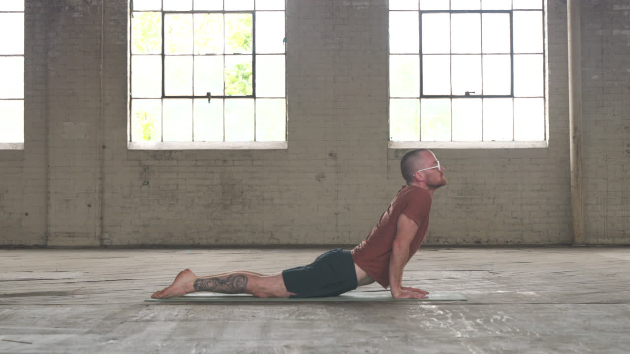Man in an industrial warehouse practicing yoga, the pose of plank to upward dog.