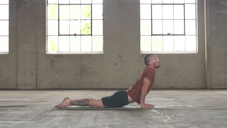 man in an industrial warehouse practicing yoga, the pose of plank to upward dog.