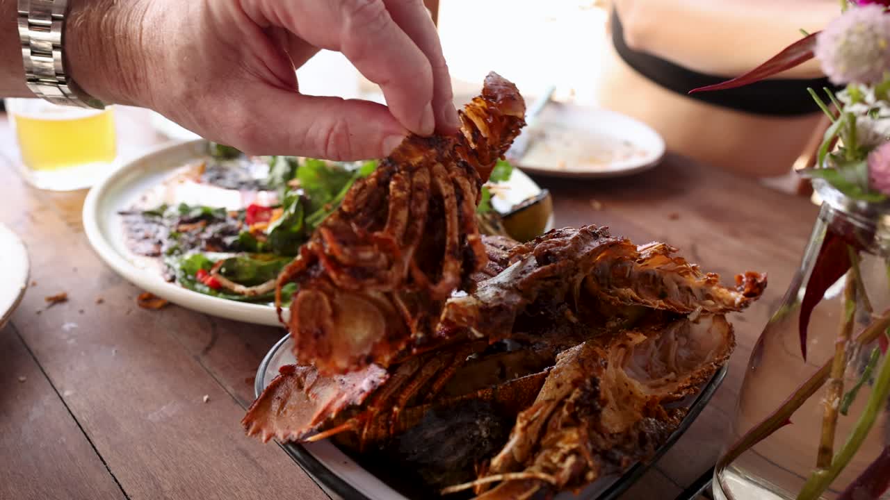 A hand serves grilled mantis shrimp on a wooden table with salad and drinks in a bright outdoor setting