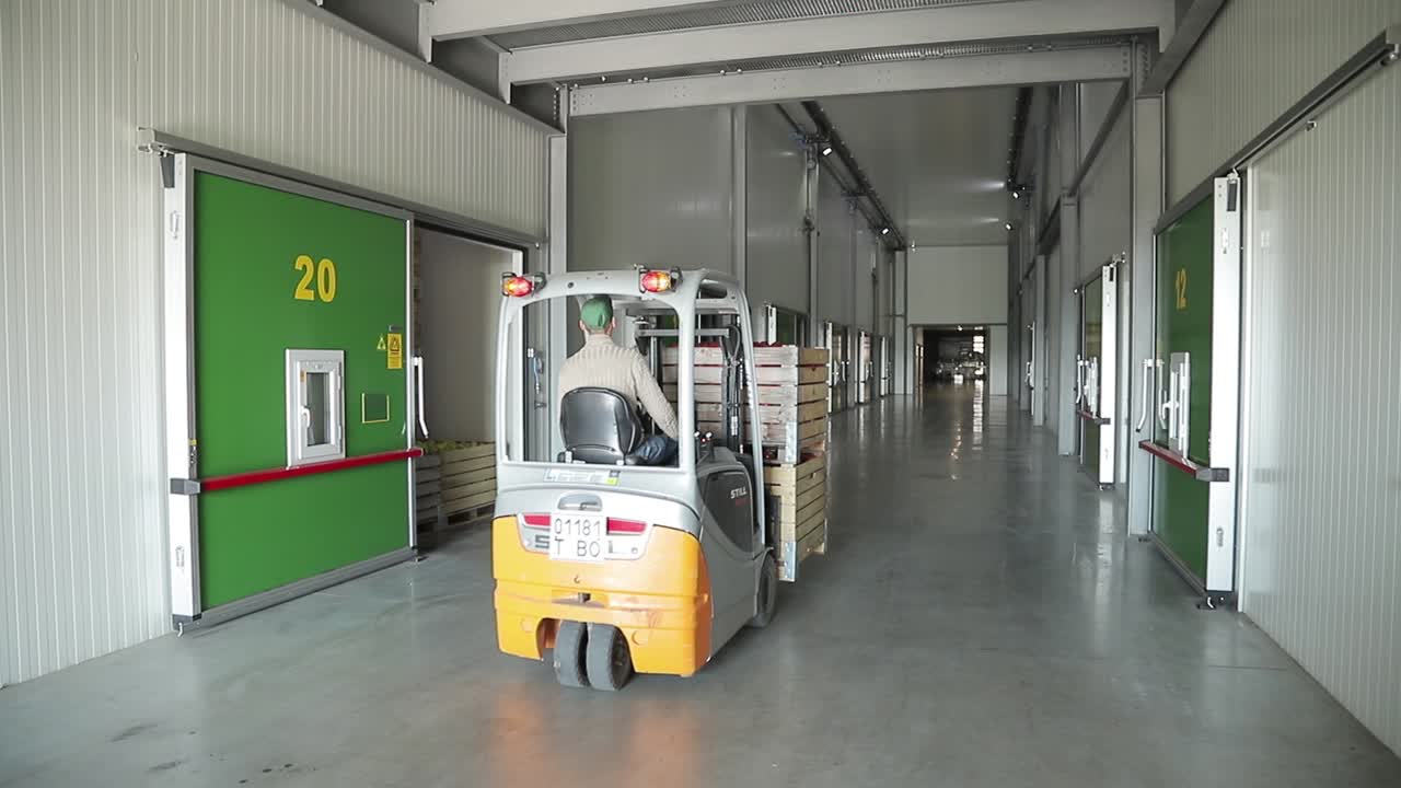A man on the loader carries two wooden boxes with red apples along the corridor for storage in the warehouse. Retail.