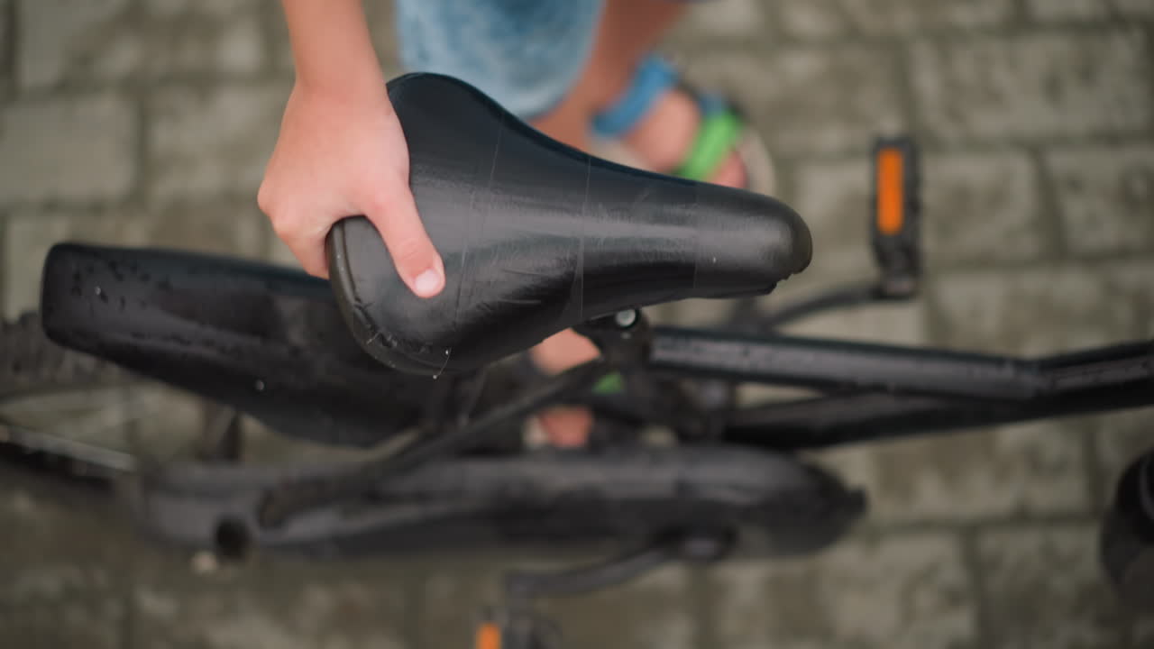 un primer plano de la mano de un niño agarrando el asiento de una bicicleta, ajustándolo cuidadosamente mientras está de pie en una superficie pavimentada húmeda, después de lo cual se aleja, el niño usa pantalones cortos de vaqueros