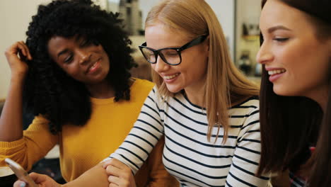 Afroamerikanische-Und-Kaukasische-Frauen,-Die-Sich-Ein-Video-Auf-Einem-Smartphone-Ansehen-Und-Kaffee-Trinken,-Sitzen-An-Einem-Tisch-In-Einem-Café