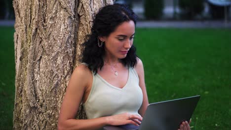 portrait of a woman in dress sits below tree at the park on grass, holding laptop on her knees, typing, doing her work