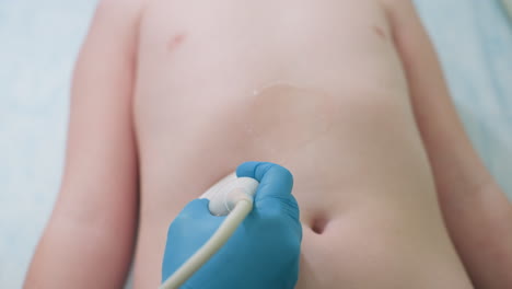 female lab attendant performs continuous ultrasound scan on upper belly of young child lying on hospital bed, using gloved hand to move probe gently across skin near diagnostic equipment