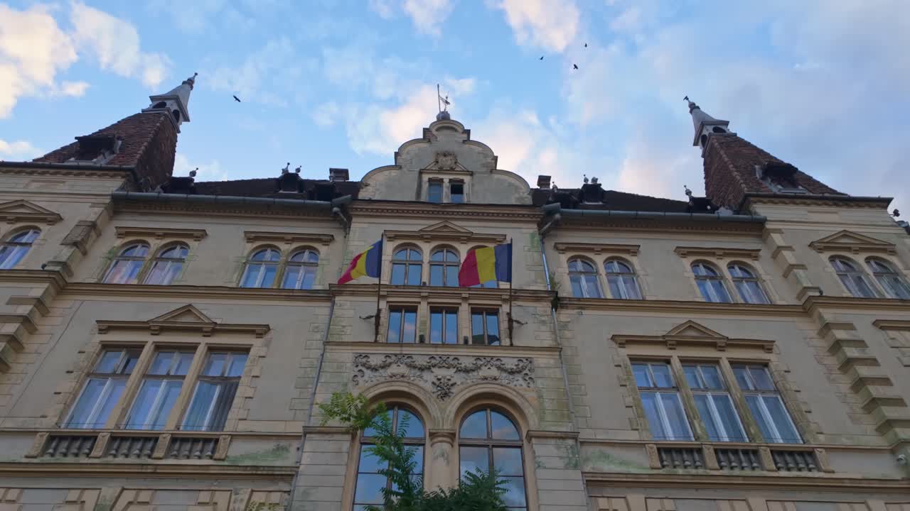 Rear facade of the Sighișoara City Hall, an elegant neo-Renaissance building with twin towers, arched windows and Romanian flags, seen under a soft evening sky