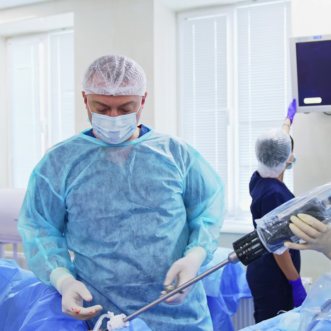 Surgical operation in process. Male doctor operates and female assistant holds the long surgical tool. Two assistants tuning the monitor at backdrop