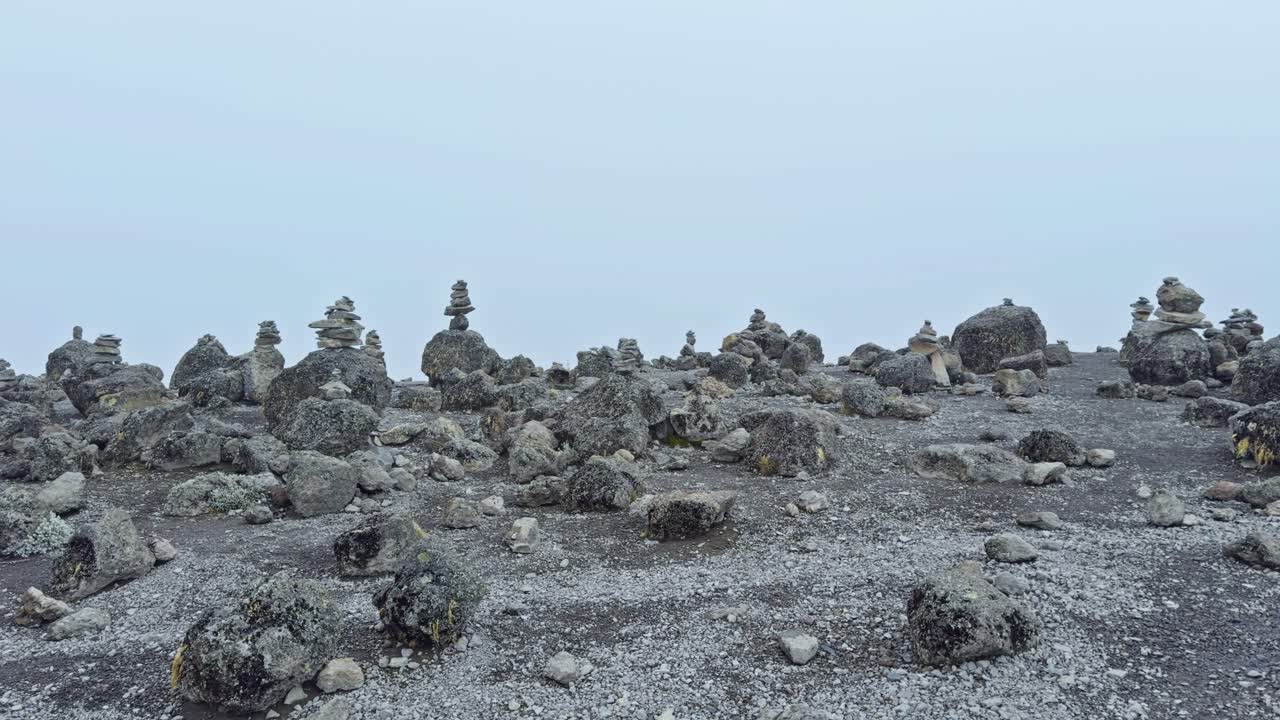 Static ground-level shot shows numerous small rock cairns scattered across a barren rocky plain at high altitude on Mount Kilimanjaro, with a pale misty sky in the background