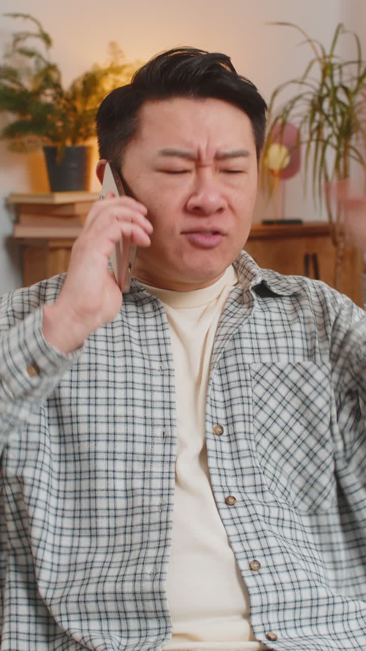 Angry young chinese man arguing on smartphone phone while sitting on couch in living room at home