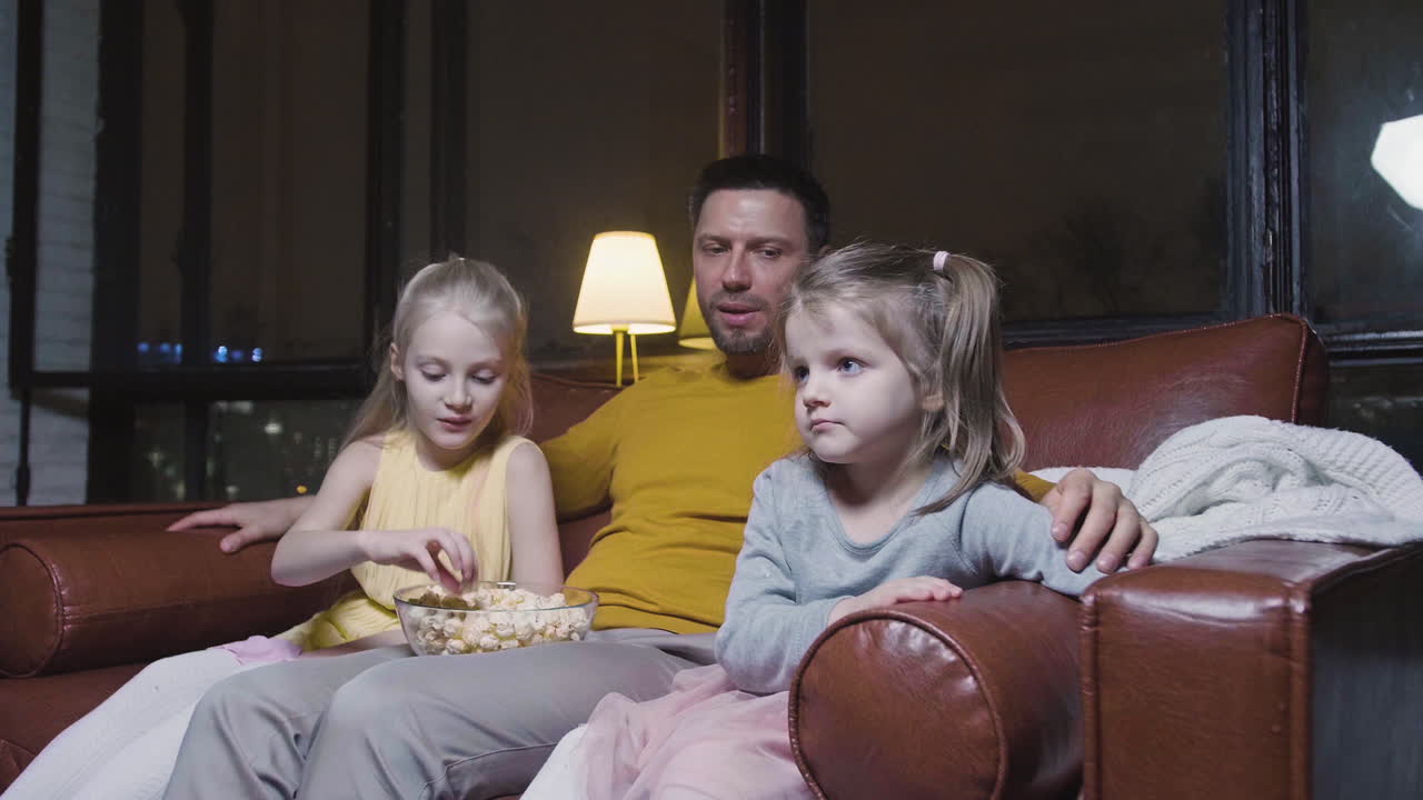 Papà e le sue due bambine che mangiano popcorn e guardano un film mentre sono sedute sul divano di notte a casa
