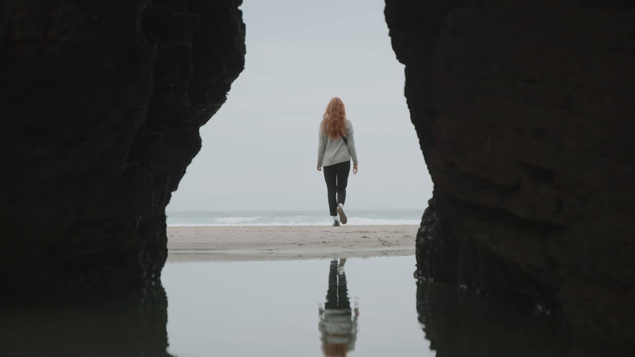 Woman walks along a secluded beach reflecting in the water, framed by a rock archway