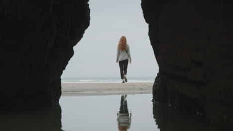 woman walks along a secluded beach reflecting in the water, framed by a rock archway