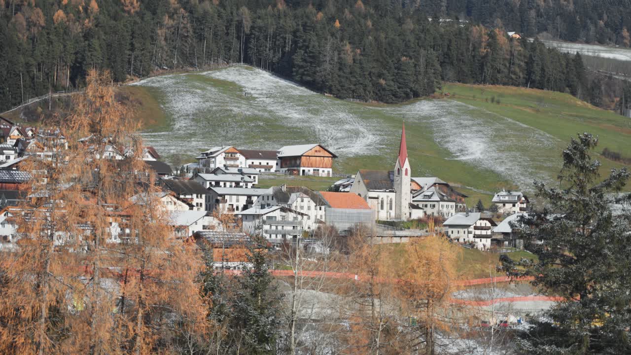 An alpine village with the red-steepled church in the center at the foot of forested hills lightly dusted with the first snow.