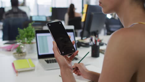 Mixed-race-businesswoman-sitting-at-desk-using-smartphone-with-copy-space