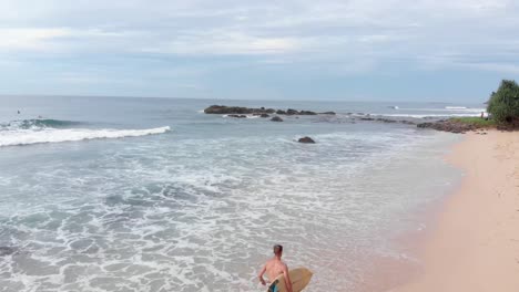 drone flying over shirtless man running on sandy beach with surfboard in sri lanka