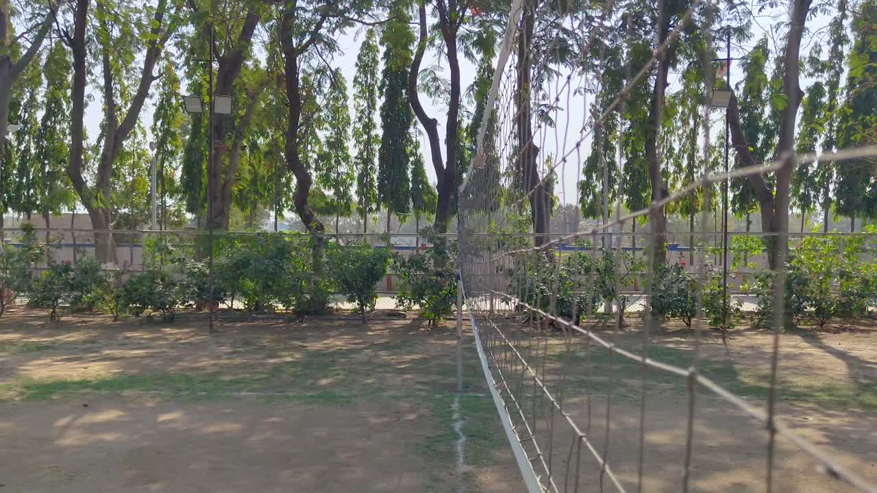View of volleyball net on outdoor court, surrounded by greenery and sunlight.