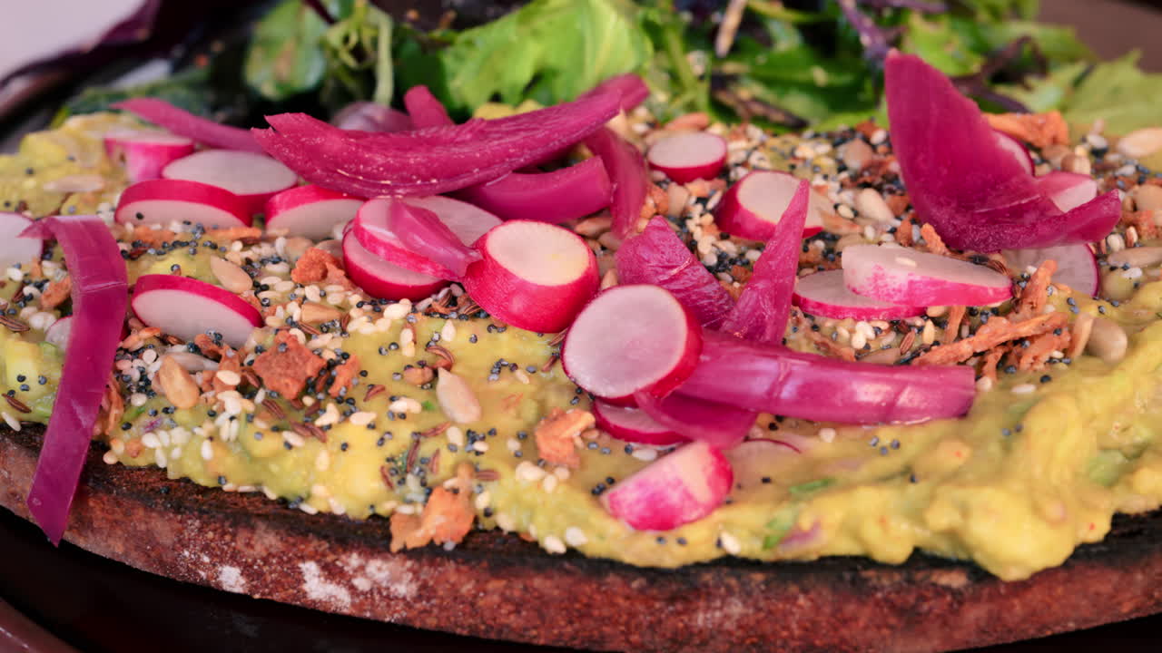 Close up of an avocado toast with seeds, radish, red onion and a side salad on a black plate