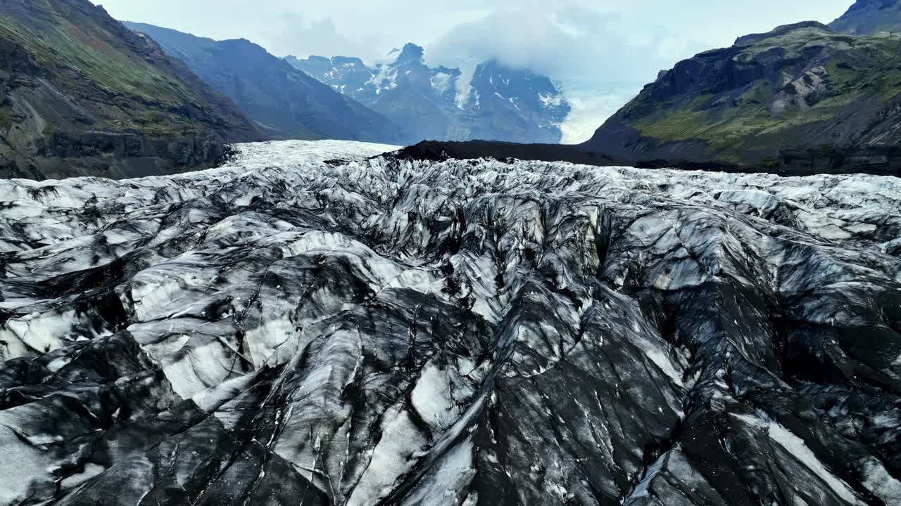 Tiefer Drohnenflug über den aschebedeckten Vatnajökull-Auslassgletscher, der tiefe Gletscherspalten und schroffe Berge unter stimmungsvollem, bewölktem Himmel in Island offenbart