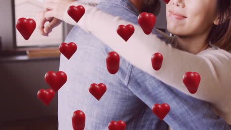 Animation-of-hearts-over-diverse-couple-embracing-and-smiling-at-home