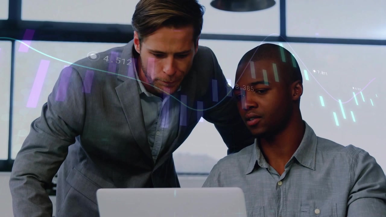 Homem sentado movendo o trackpad, iniciando barras e linhas financeiras sobrepostas no laptop, com um homem em pé orientando.