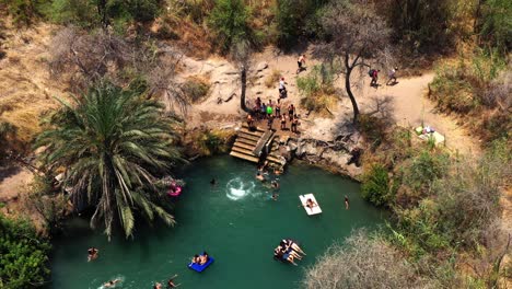 many people float on air mattresses on a warm sun-drenched day on the clear blue natural spring while young people enthusiastically splash from a platform into the water