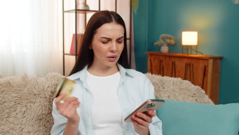young woman sitting on sofa anxiously checking credit card balance on smartphone worried about error