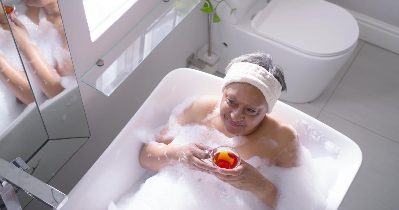 Senior African American woman reclining in bath, looking at clear cup and sipping for relaxation