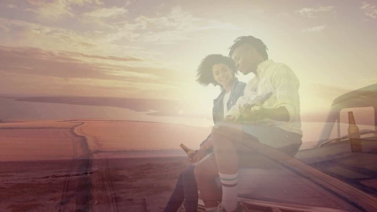 Animation of african american couple sitting on car drinking beers at beach over sunset landscape