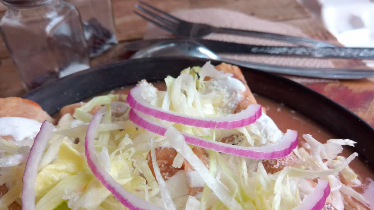 Zoom in on a plate of enchiladas topped with lettuce, cream, and red onions. Traditional Mexican dish presented on a rustic wood surface.